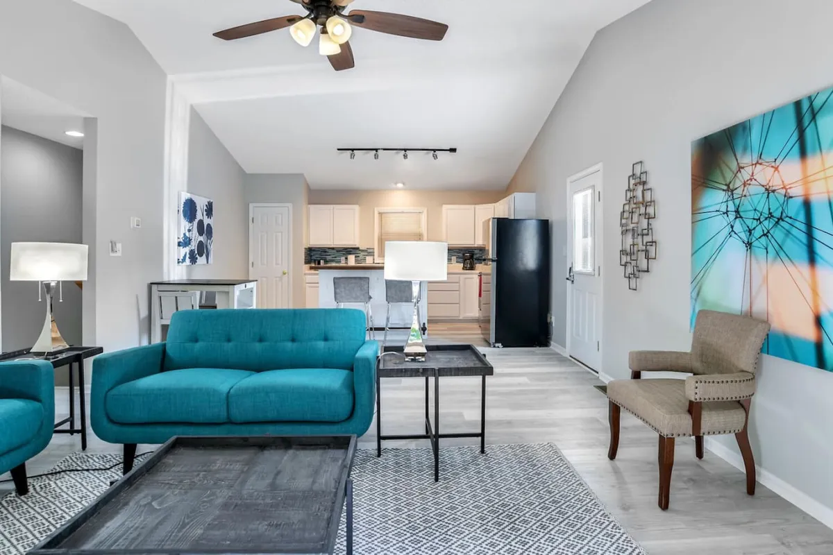 An open-concept living area featuring teal sofas, modern kitchen appliances, wall art, and stylish lighting. The layout suggests seamless movement from the entryway through social and kitchen spaces, with the front door likely nearby.