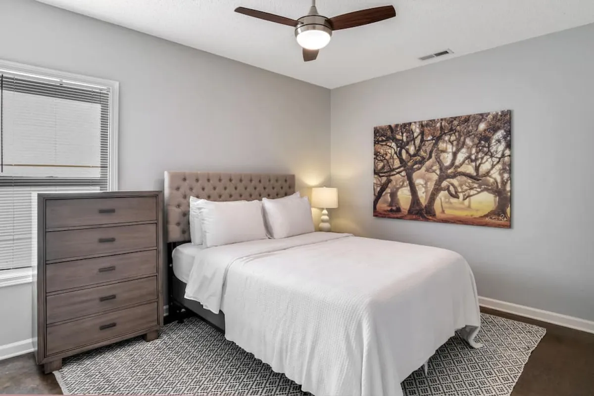 A cozy bedroom with a tufted headboard, soft bedding, artwork featuring trees, a nightstand with a lamp, and a dresser. The design is modern yet inviting, with an emphasis on comfort and relaxation.