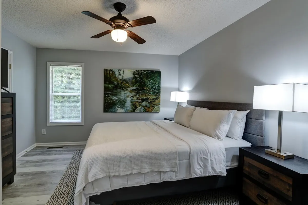 A bedroom featuring a comfortable bed with soft linens, complemented by tasteful artwork, two nightstands, and a window that lets in gentle natural light. The space is uncluttered, with clean lines and a serene color palette.