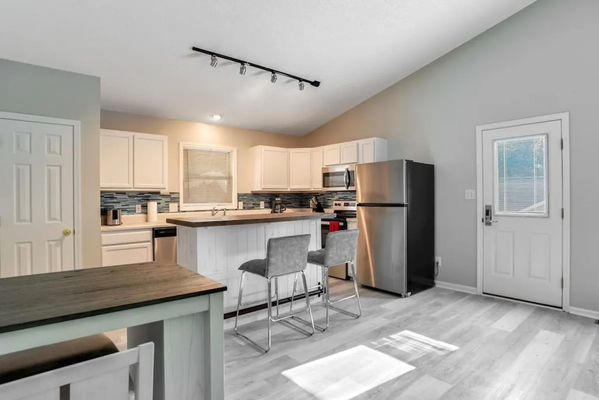 A bright, welcoming kitchen with a central island, stainless steel appliances, and bar stools. Natural light pours in through a nearby door, suggesting proximity to a ground-level entrance or garage for easy grocery unloading.