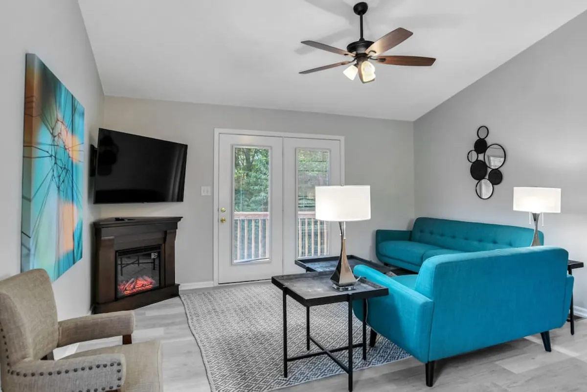 The image showcases a stylish, contemporary living room with a vibrant turquoise couch as the centerpiece. Abstract wall art adds a modern touch above the couch, while a sleek fireplace and flat-screen television create a cozy entertainment zone. Large windows let in natural light, enhancing the inviting atmosphere, perfect for relaxing with loved ones or entertaining friends.