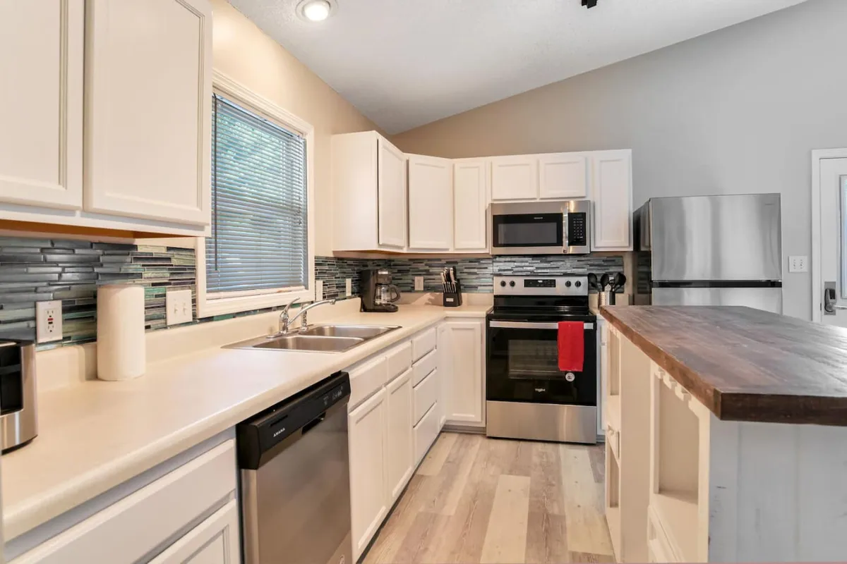 A modern kitchen space featuring white cabinetry, a warm wooden countertop, stainless steel appliances, and a glass tile backsplash. The layout is inviting for cooking together and hosting, with thoughtful design details that add a contemporary touch.
