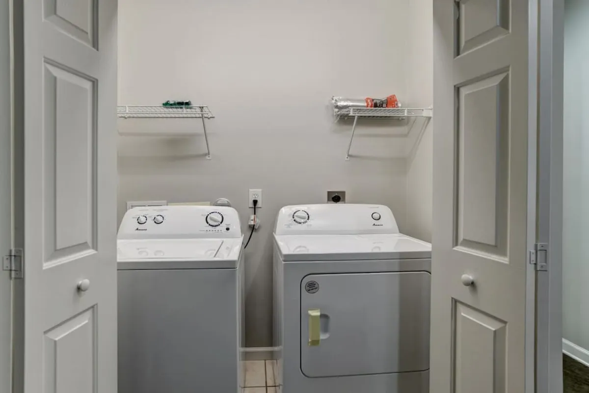 A compact laundry room with a modern washing machine and drying machine, open shelving above for cleaning supplies and laundry essentials. The space is organized and functional, positioned near a ground-floor entryway, hinting at immediate access and practical everyday living.