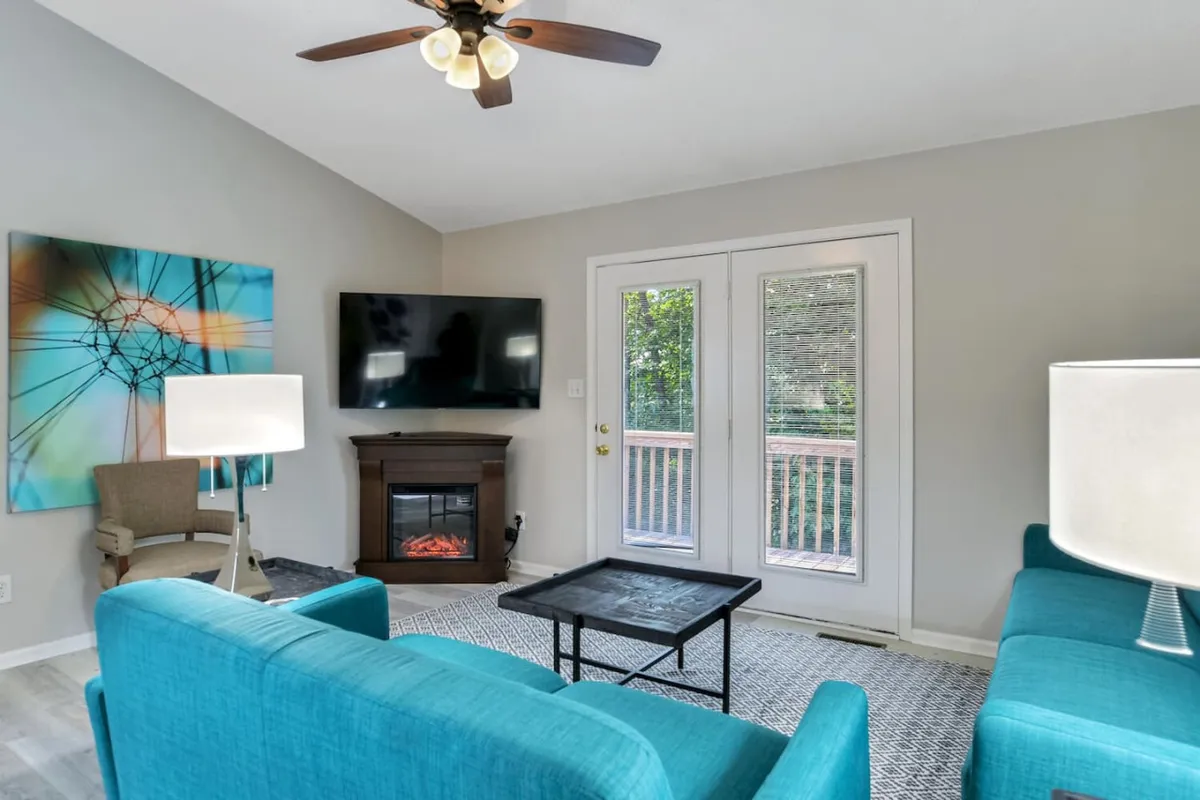 A comfortable, modern living room with a vibrant turquoise sofa, bold artwork, fireplace, and large sliding glass doors letting in morning sunlight. The setup feels perfect for entertaining or relaxing, with easy access to the outdoors.