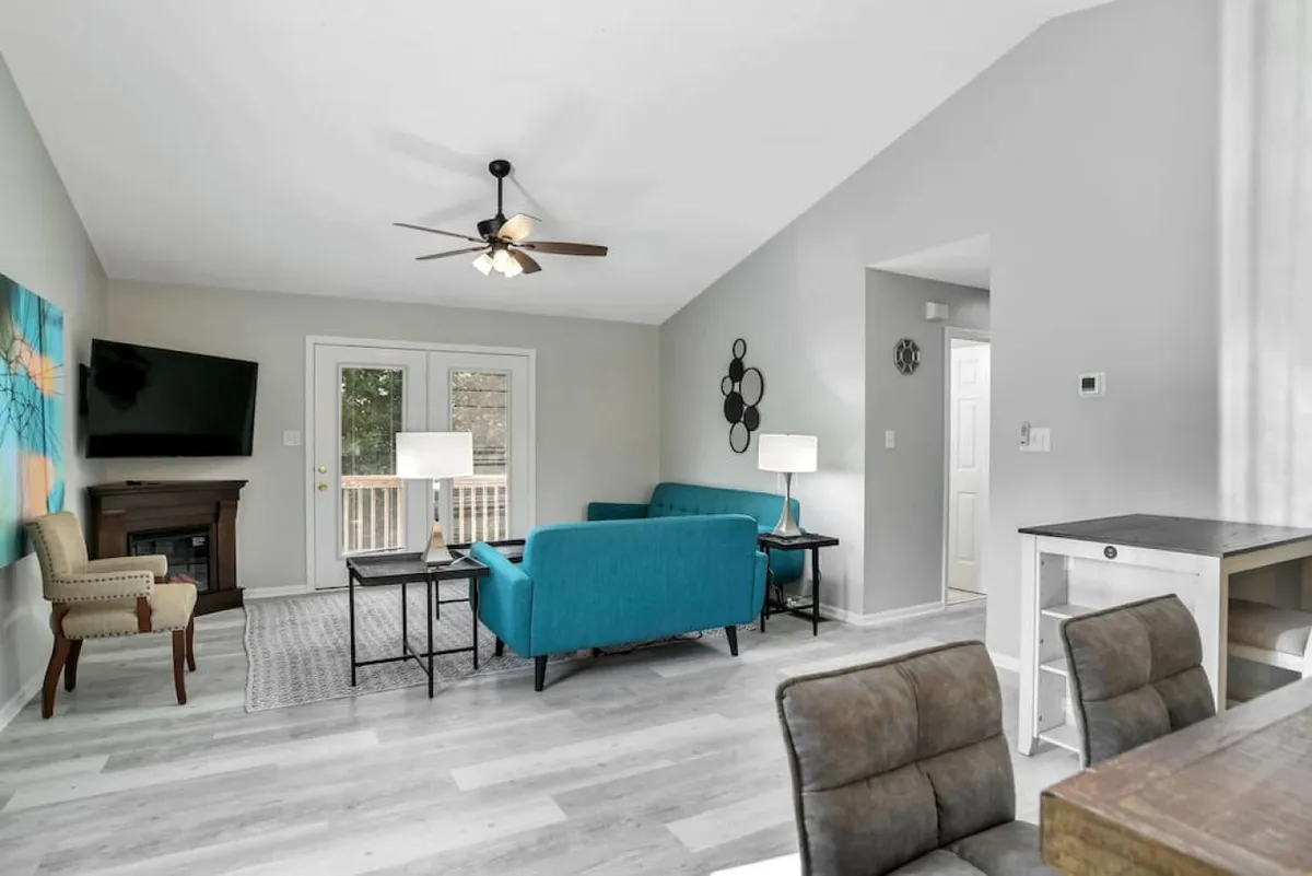 A welcoming living room with turquoise couches, a wooden coffee table, large windows letting in abundant natural light, and a modern light fixture. The arrangement of seating encourages conversation, while the windows reveal views of the deck and trees outside.